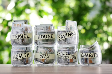 Glass jars with money for different needs on table against blurred background