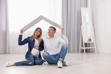 Photo of Young family housing concept. Pregnant woman with her husband sitting under cardboard roof on floor indoors. Space for text