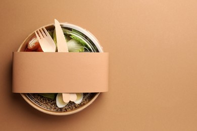 Photo of Tasty food in container with wooden fork and knife on beige background, top view. Space for text