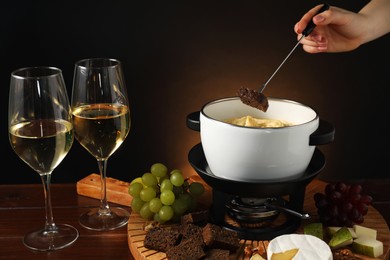 Photo of Woman dipping piece of bread into fondue pot with melted cheese at wooden table with wine and snacks, closeup