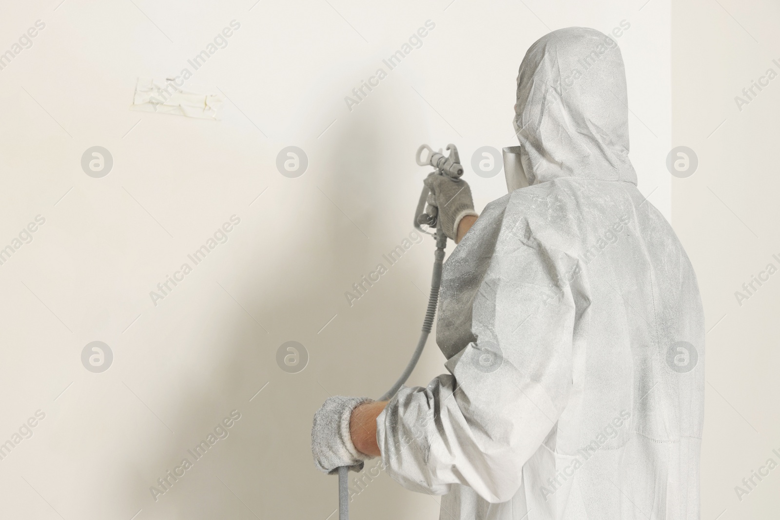 Photo of Decorator in protective overalls painting wall with spray gun indoors