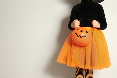 Photo of Cute little girl with pumpkin candy bucket wearing Halloween costume on light background, closeup. Space for text