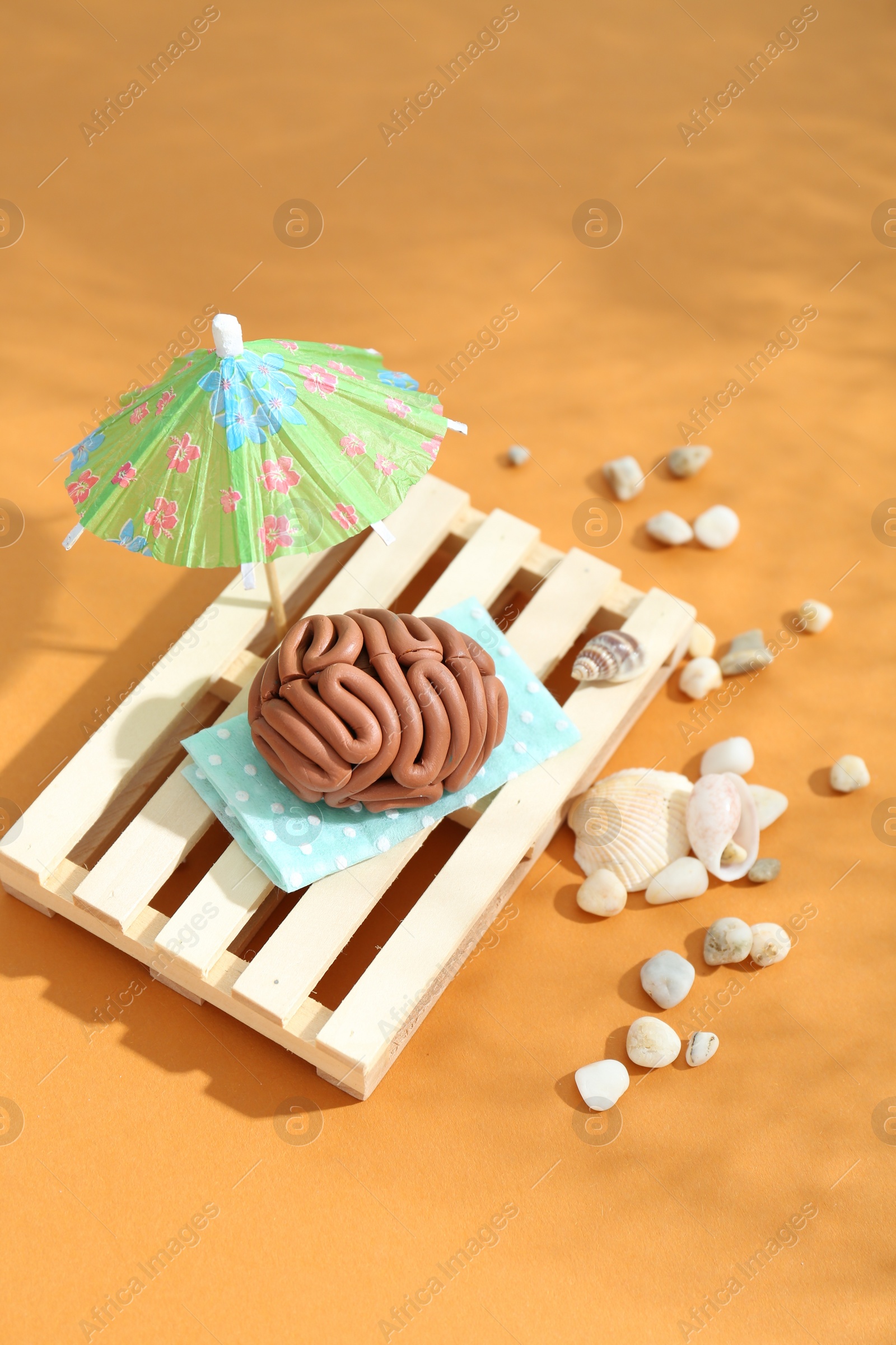 Photo of Brain made of plasticine on mini wooden sunbed under umbrella against orange background
