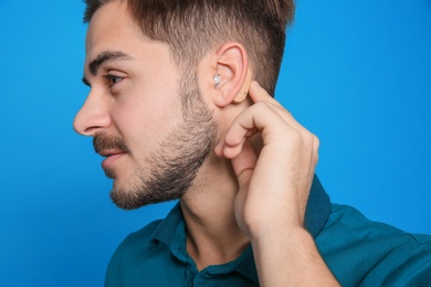 Young man adjusting hearing aid on color background