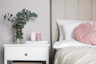Photo of Vase with beautiful eucalyptus branches, book and candles on nightstand in bedroom
