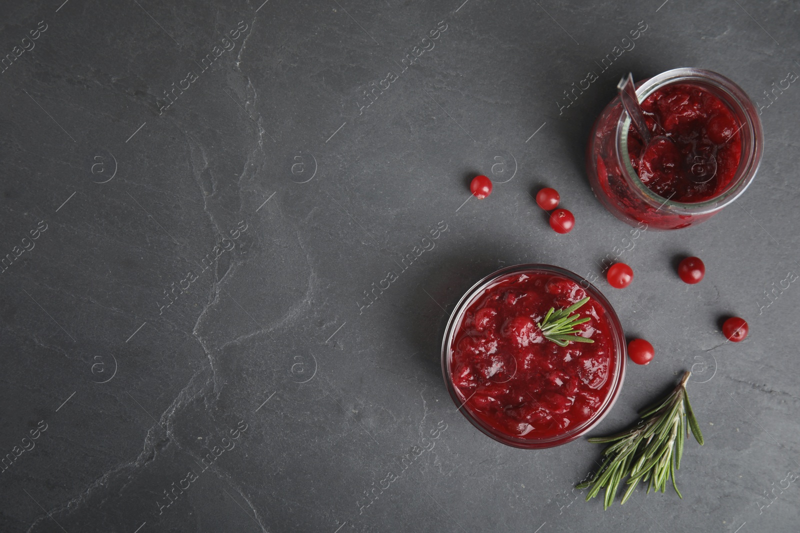 Photo of Jar and bowl of cranberry sauce with rosemary on grey background, flat lay. Space for text