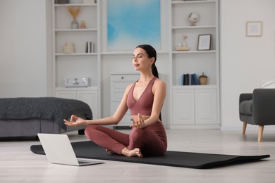 Beautiful young woman practicing Padmasana with laptop on yoga mat at home. Lotus pose