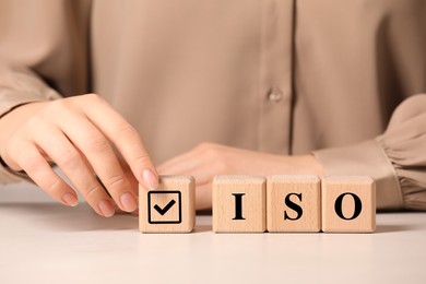 Photo of Woman adding cube with check mark to abbreviation ISO of wooden cubes at white table, closeup