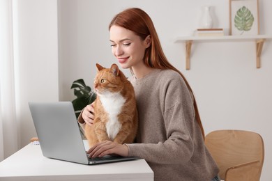 Photo of Woman working with laptop and hugging cat at desk. Home office