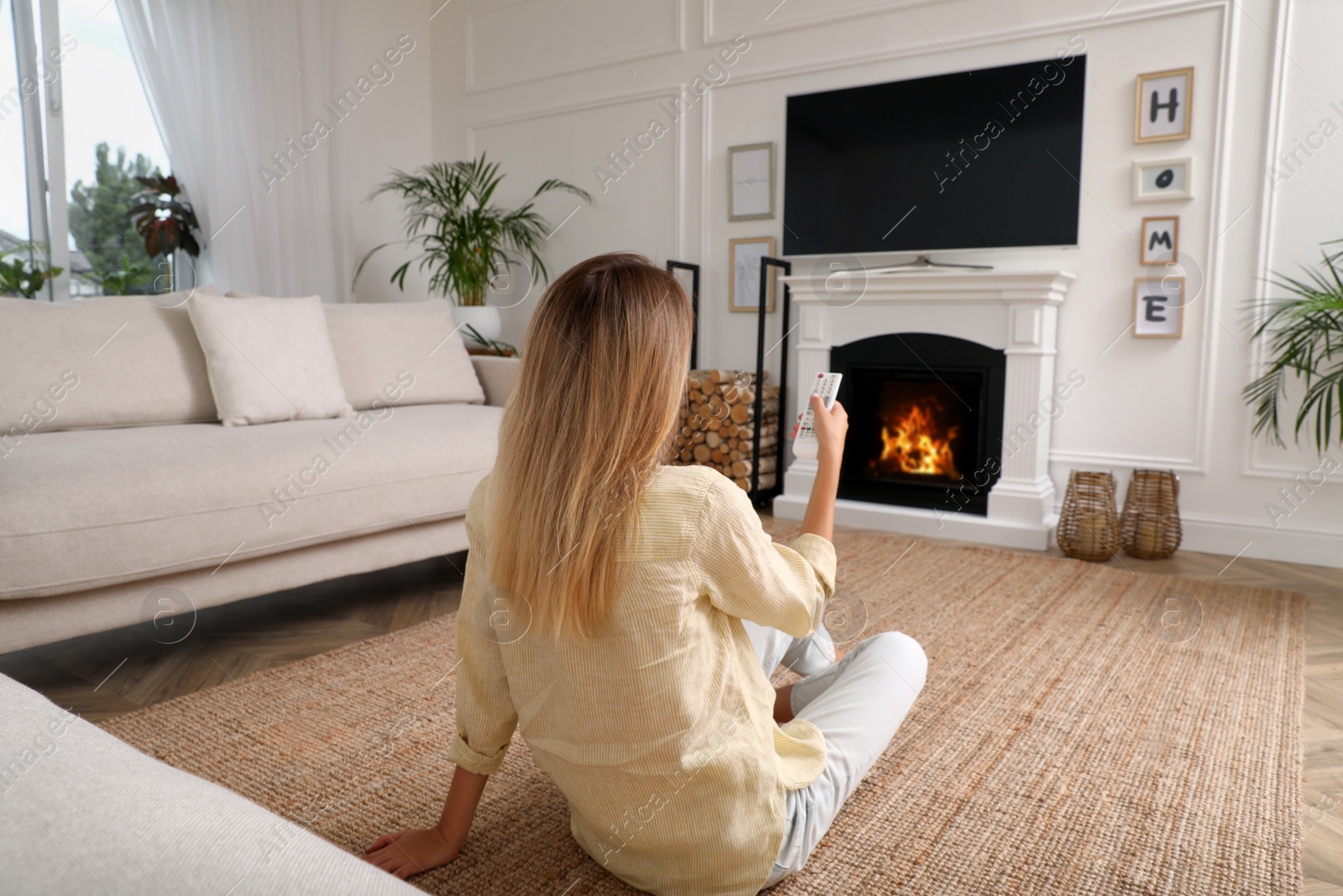 Photo of Young woman watching television at home, back view. Living room interior with TV on fireplace