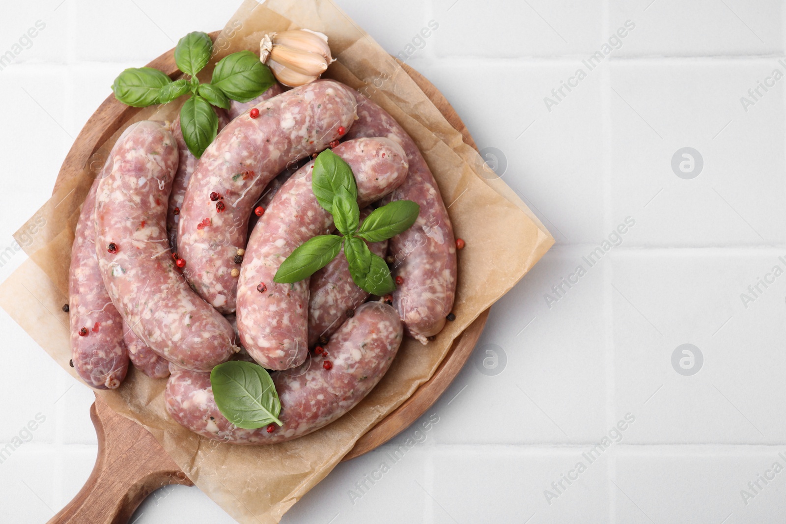 Photo of Board with raw homemade sausages, basil leaves and peppercorns on white tiled table, top view. Space for text