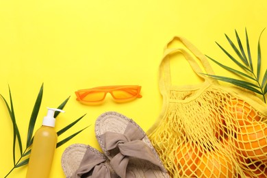 String bag with oranges, sunglasses, cosmetic product, slippers and palm leaves on yellow background, flat lay