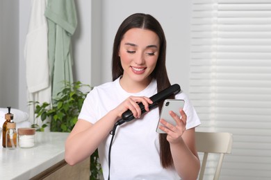 Photo of Beautiful young woman with hair iron using smartphone in bathroom. Internet addiction