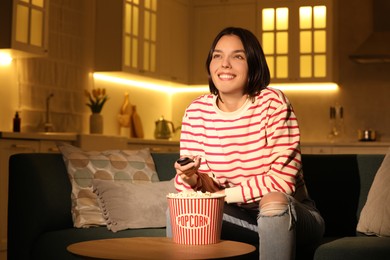 Happy woman watching TV with popcorn on sofa at home, space for text