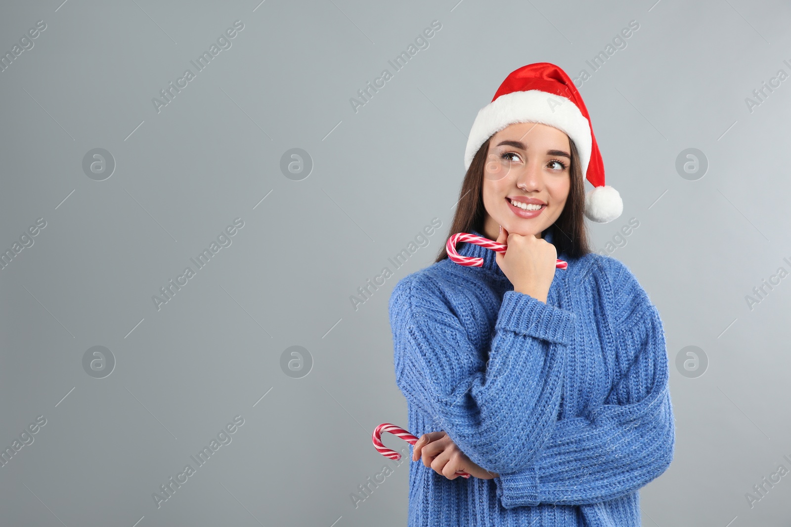 Photo of Young woman in blue sweater and Santa hat holding candy canes on grey background, space for text. Celebrating Christmas
