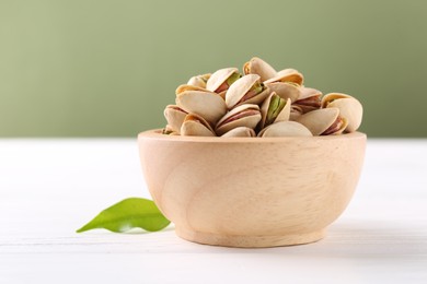 Tasty pistachios in bowl on white wooden table against olive background, closeup