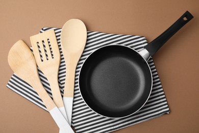 Different kitchen utensils, napkin and frying pan on light brown background, flat lay