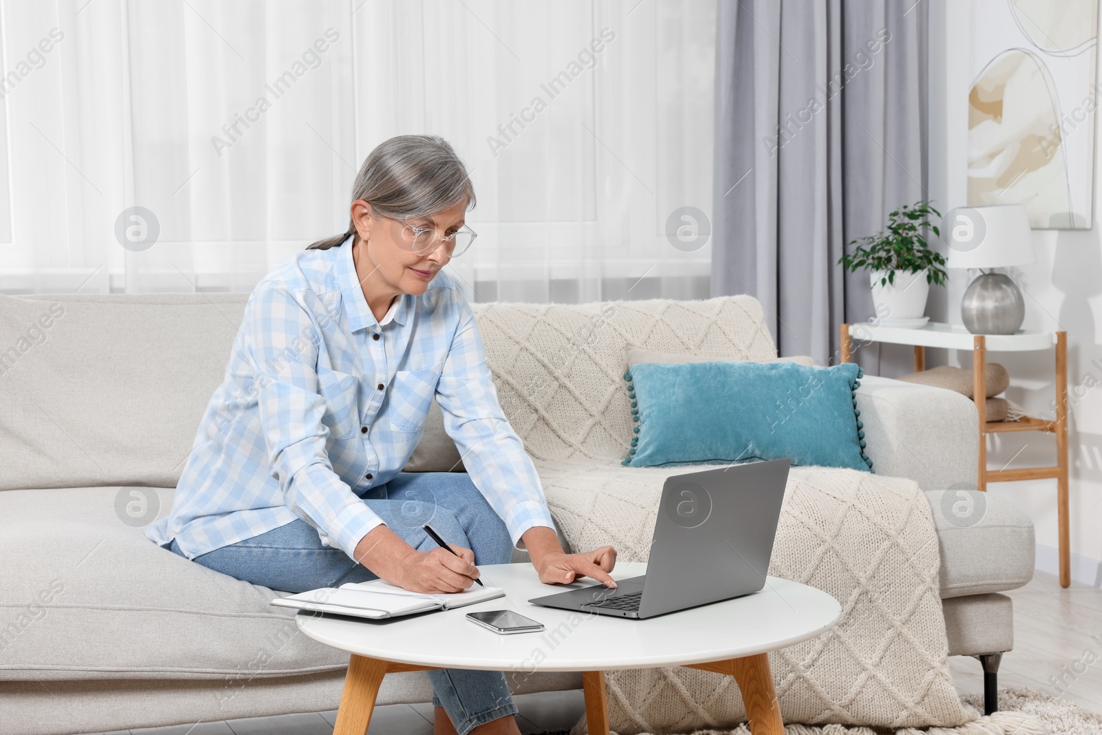 Photo of Beautiful senior woman writing something in notebook while using laptop at home