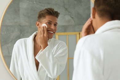 Handsome young man cleaning face with cotton pad near mirror in bathroom