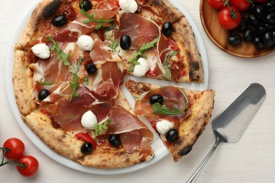 Tasty pizza with cured ham, olives, mozzarella cheese, tomatoes and arugula on light wooden table, flat lay