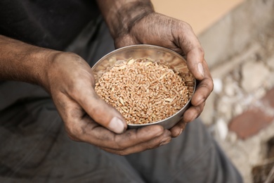 Poor homeless man with bowl of wheat outdoors, closeup