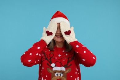 Senior woman in Christmas sweater and Santa hat covering face with hands in knitted mittens on light blue background