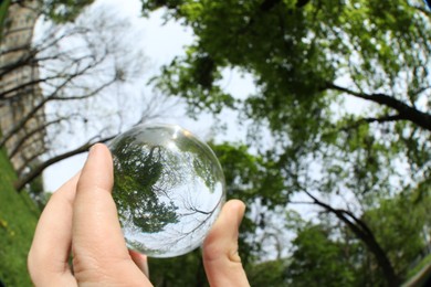 Photo of Beautiful green trees outdoors, overturned reflection. Man holding crystal ball in park, closeup. Wide-angle lens