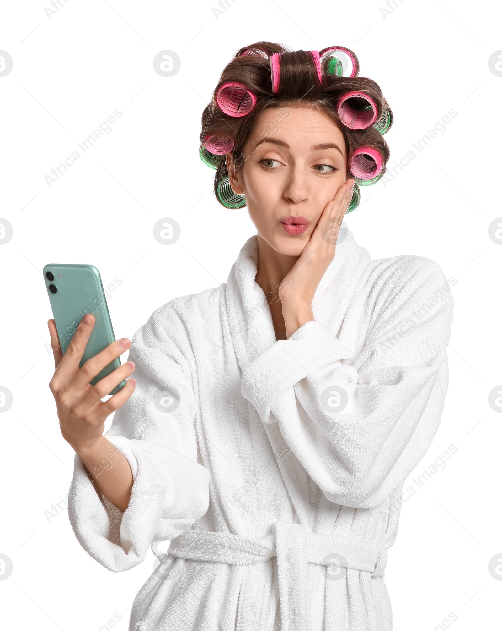 Photo of Emotional young woman in bathrobe with hair curlers taking selfie on white background