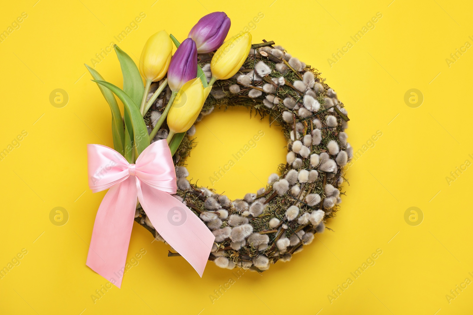 Photo of Wreath made of beautiful willow, colorful tulip flowers and pink bow on yellow background, top view