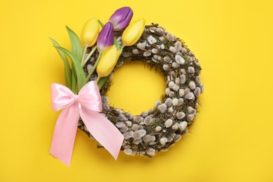 Photo of Wreath made of beautiful willow, colorful tulip flowers and pink bow on yellow background, top view