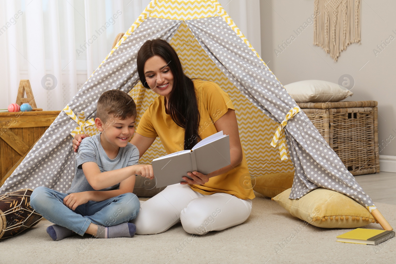 Photo of Mother and son reading book near toy wigwam at home