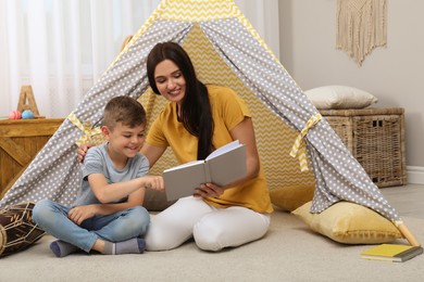 Photo of Mother and son reading book near toy wigwam at home