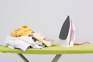 Photo of Board with modern iron and pile of clothes on light background. Laundry day