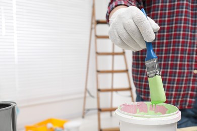 Man dipping brush into bucket of green paint indoors, closeup. Space for text