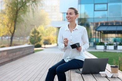 Photo of Smiling businesswoman with lunch box sitting near laptop and paper cup outdoors. Space for text