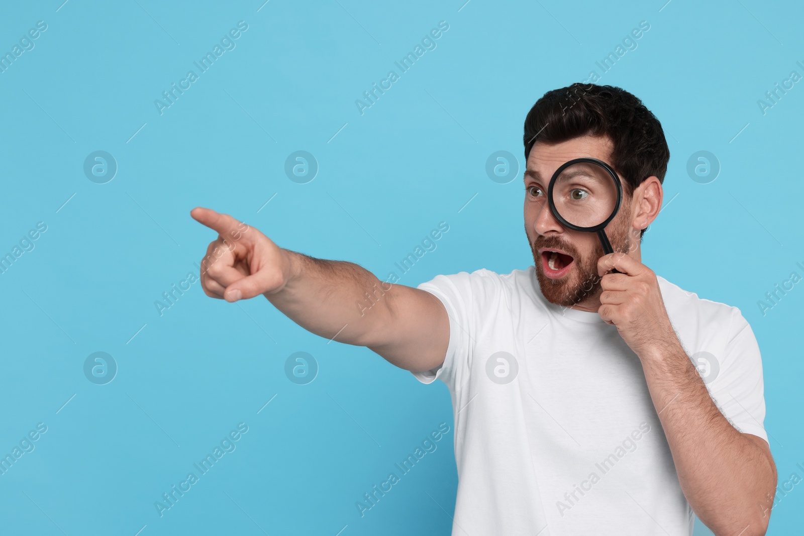 Photo of Man looking through magnifier glass and pointing on light blue background, space for text