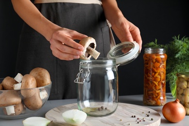 Photo of Woman putting champignon into glass jar at grey table, closeup. Pickling mushrooms