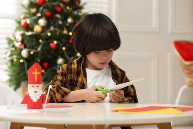 Photo of Cute little boy cutting paper at table with Saint Nicholas toy indoors