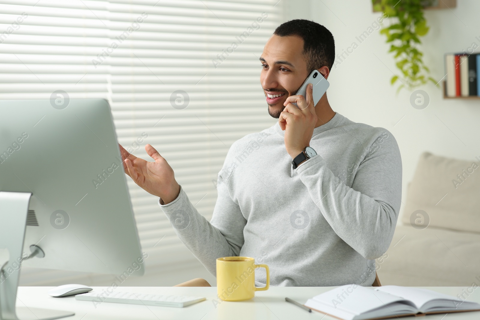 Photo of Young man talking on smartphone while working with computer at desk in room. Home office