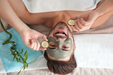 Photo of Young man with clay mask on his face holding cucumber slices in spa salon, above view