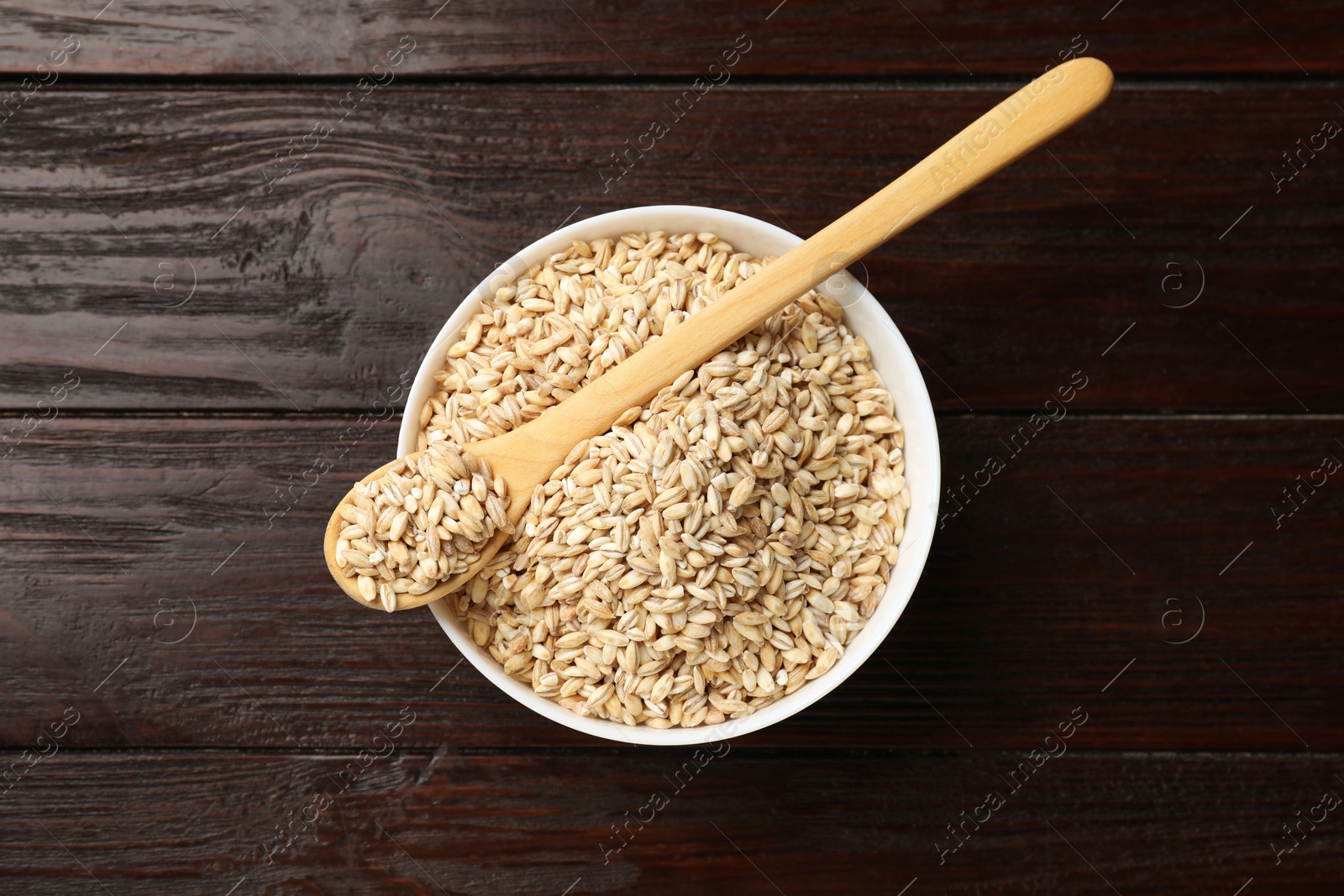 Photo of Dry pearl barley in bowl and spoon on wooden table, top view