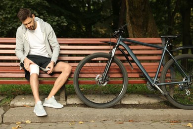 Photo of Man applying bandage onto his knee on wooden bench outdoors