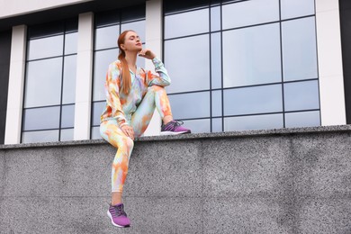 Beautiful woman in gym clothes sitting on parapet on street, space for text