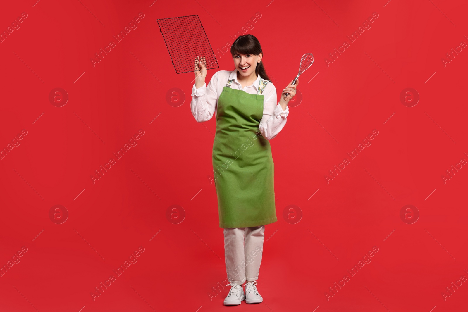 Photo of Happy confectioner in apron holding professional whisk and cooling rack on red background