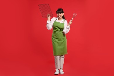 Photo of Happy confectioner in apron holding professional whisk and cooling rack on red background