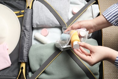 Woman putting small bottle with personal care product into plastic bag near suitcase, closeup. Cosmetic travel kit