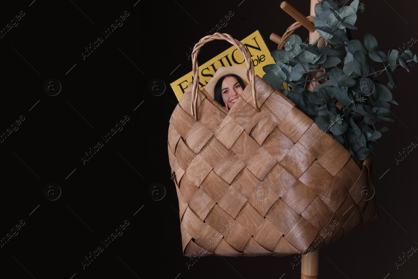 Photo of Stylish wicker basket with green plant and magazine hanging on clothing rack against dark background, space for text