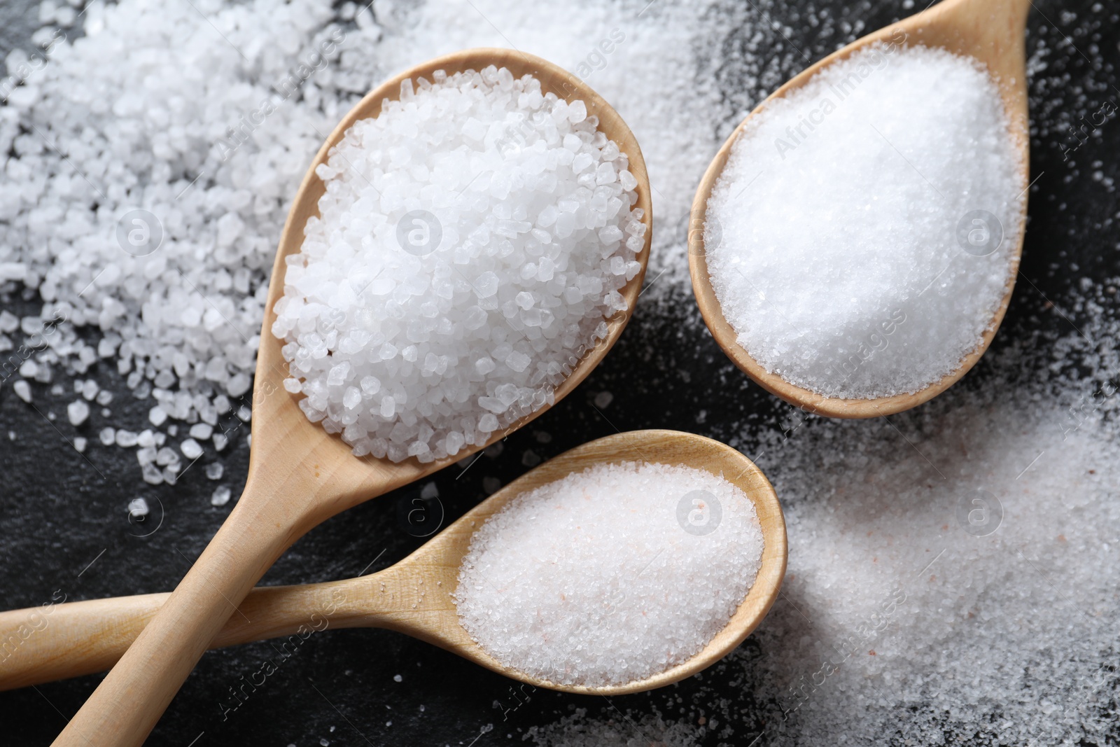 Photo of Organic salt in spoons on black table, flat lay