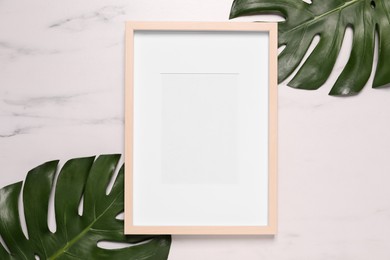 Photo of Empty photo frame and green leaves on white marble background, flat lay. Space for design