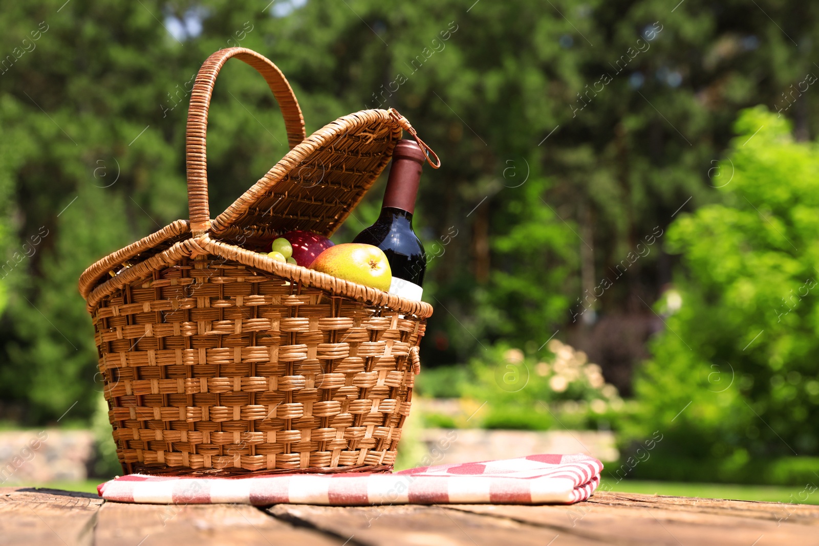 Photo of Picnic basket with fruits, bottle of wine and checkered blanket on wooden table in garden